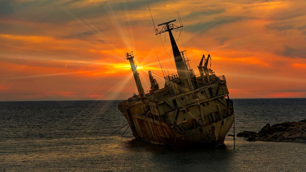 Rusted cargo shipwreck listing near a rocky shore with a sunburst at sunset.
