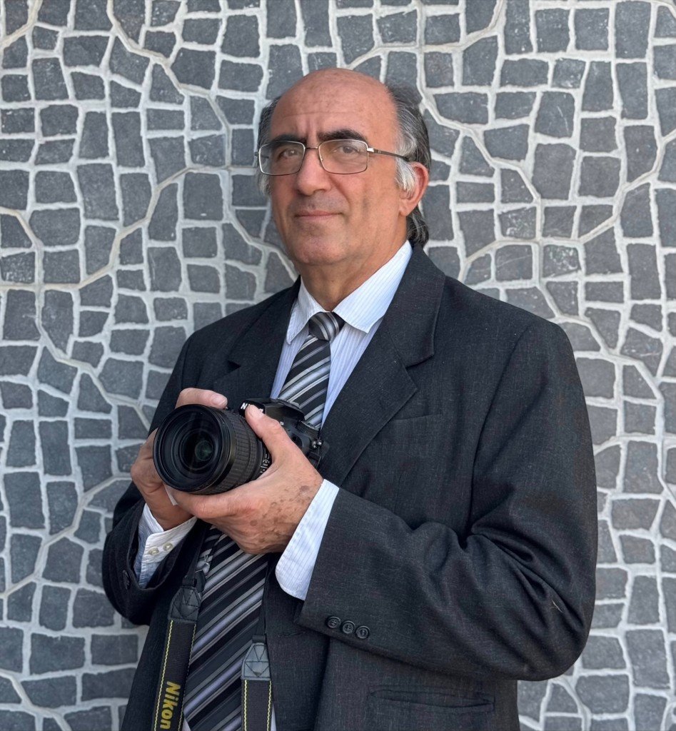 Panicos Stelikos holding a Nikon camera, wearing a dark suit and glasses.